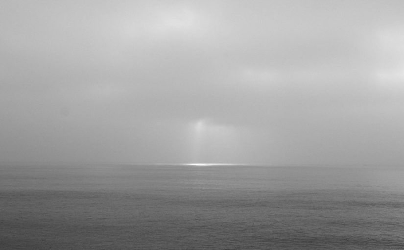 Black and white seascape photograph with sun's rays shining through clouds on the horizon